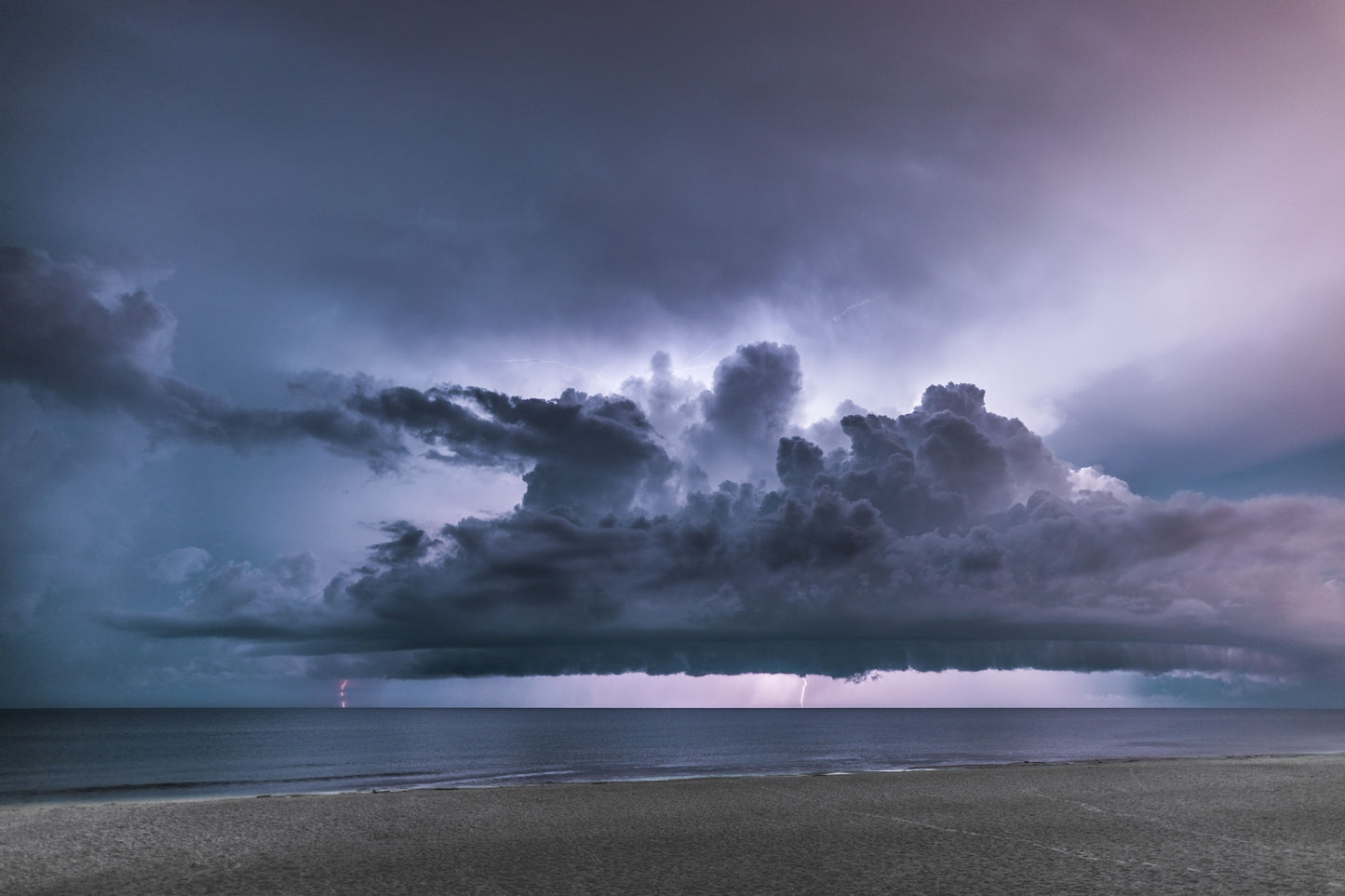 A wide violet-lit storm cloud stretching over a calm Gulf sea at dusk with lightning striking the horizon above an empty beach — fine art coastal storm photography by Susan Gunn.