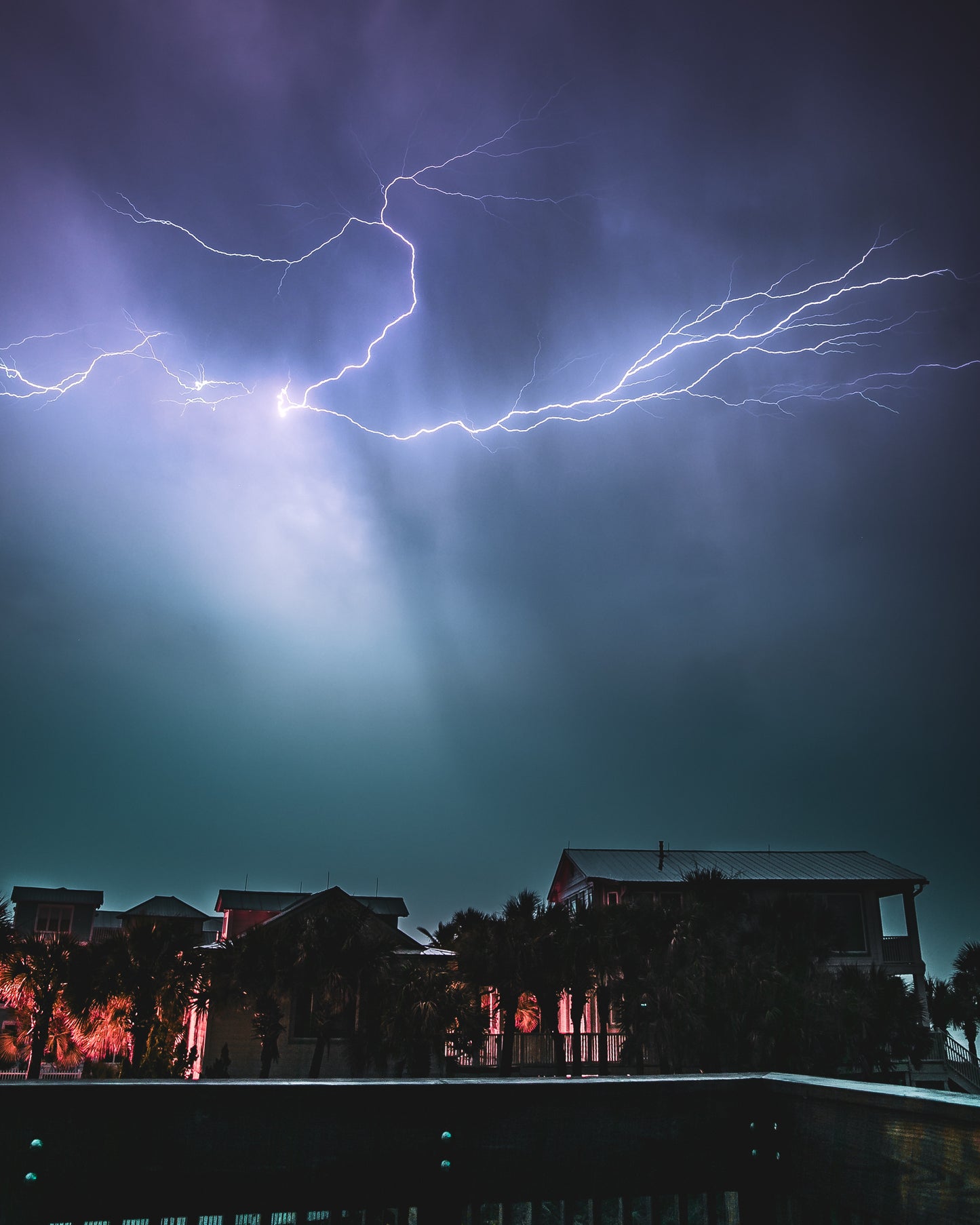Spider lightning branching across a night sky over silhouetted Gulf Coast beach homes with red-lit palms, a glowing rain shaft below — fine art storm photography by Susan Gunn.