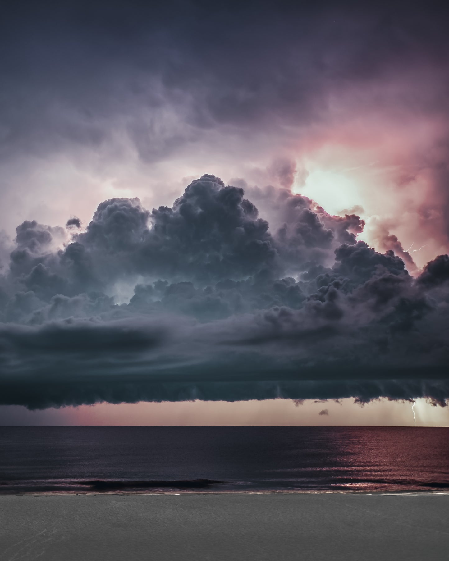 A towering pink-lit thunderhead crowning over a calm Gulf sea and pale beach at dusk, with distant lightning — fine art storm photography by Susan Gunn.