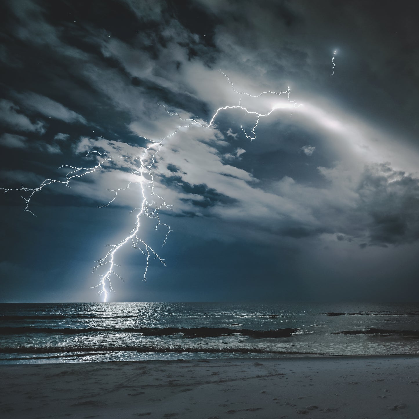 A lightning bolt strikes the Gulf at night as waves glow silver over white sand — fine art storm photography by Susan Gunn.