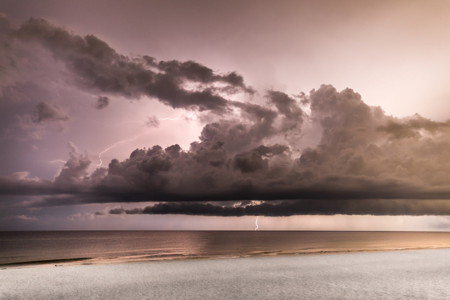 A pastel dusk storm over the Gulf — calm sea and white-sand beach below dramatic clouds, with lightning striking the horizon — fine art coastal photography by Susan Gunn.