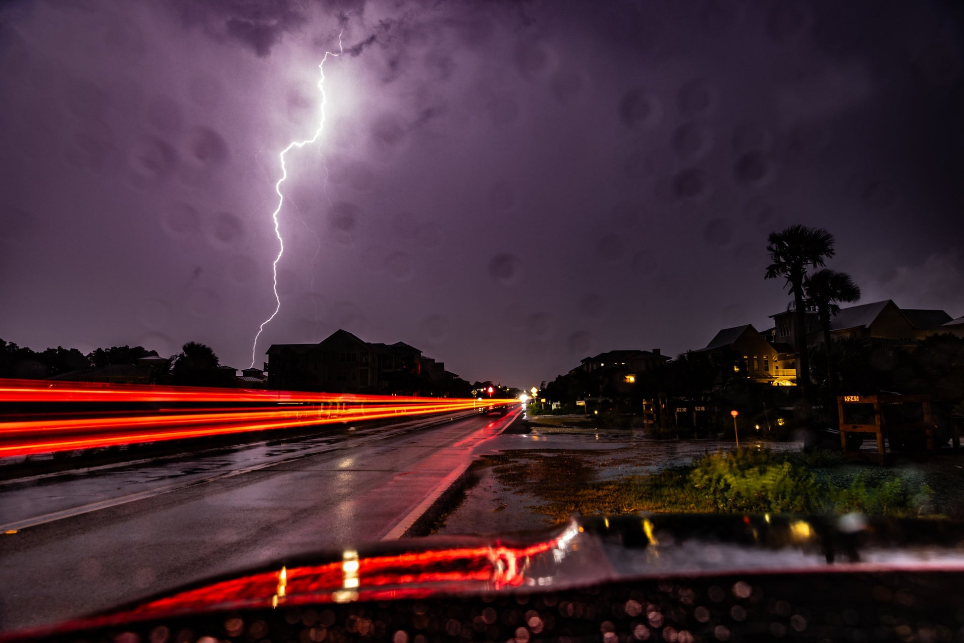 Lightning striking over coastal rooftops at night during a storm, with red car-light trails streaking down a wet road — fine art photography by Susan Gunn.