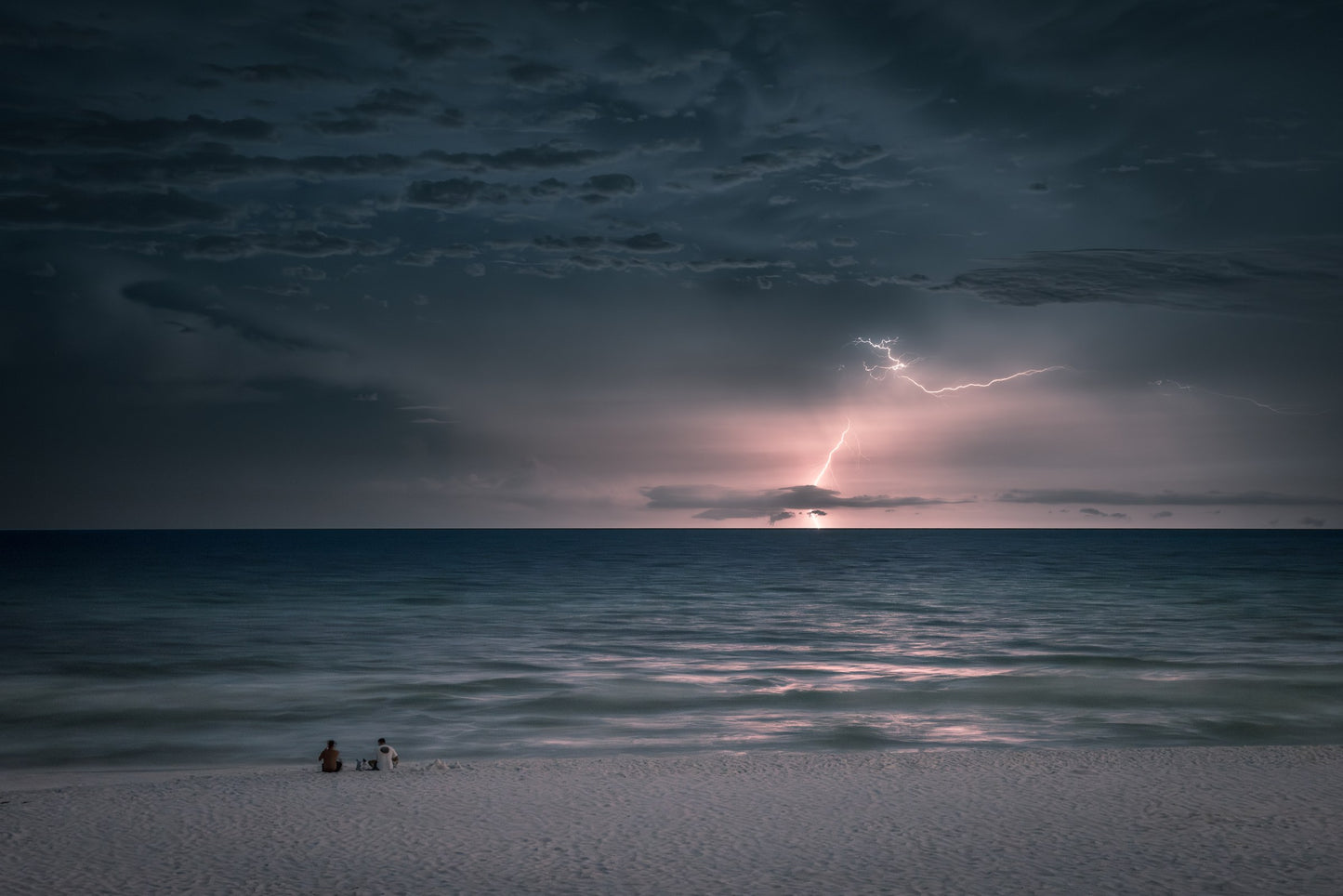 Two people sit on a white-sand Gulf beach at dusk looking at their phones while distant lightning strikes the horizon over a calm sea — fine art coastal photography by Susan Gunn.